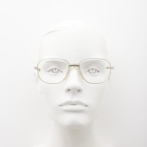 Puede incluir: Gafas de metal en tono dorado con montura rectangular de esquinas redondeadas. Las gafas tienen un puente fino y patillas delgadas, con lentes transparentes colocadas sobre una cabeza de maniquí blanca sobre un fondo liso.