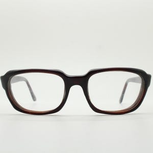 Puede incluir: Gafas marrones oscuras con montura rectangular. Las gafas tienen una montura gruesa y redondeada y lentes transparentes. Las patillas son finas y marrones oscuras. Fondo blanco.