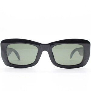 Può includere: Occhiali da sole rettangolari con montatura in plastica nera lucida e lenti color verde scuro. L'occhiale presenta un ponte spesso e solido e aste larghe che si curvano verso l'interno, offrendo un aspetto audace, strutturato e ben definito.