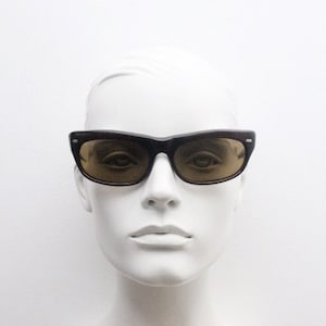 May include: Black-framed sunglasses with brown-tinted lenses. The sunglasses have a classic, rectangular shape and are displayed on a white mannequin head. The lenses reflect the surrounding environment.