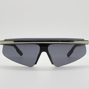 Puede incluir: Gafas de sol negras con una banda de metal plateado en la parte superior. Las lentes son de color gris oscuro y la montura es angular.