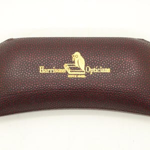 Puede incluir: Un estuche para gafas texturizado de color burdeos oscuro con forma rectangular curva. En el centro, el texto dorado "Harrisons Opticians" aparece junto a un icono de un búho con gafas sobre un libro encima de las palabras "ESTD 1908."