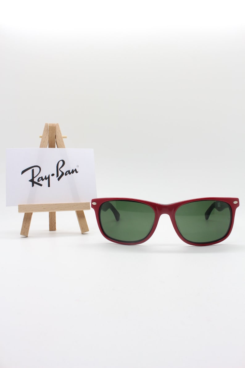 May include: Red Ray-Ban sunglasses with green lenses on a white background. The sunglasses are sitting on a small wooden easel with a white card that says "Ray-Ban" on it.