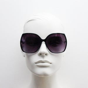 Puede incluir: Gafas de sol negras de gran tama&ntilde;o con lentes de color morado oscuro. Las gafas est&aacute;n en una cabeza de maniqu&iacute; blanca.