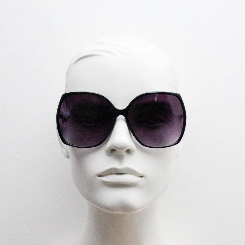 Puede incluir: Gafas de sol negras de gran tama&ntilde;o con lentes de color morado oscuro. Las gafas est&aacute;n en una cabeza de maniqu&iacute; blanca.
