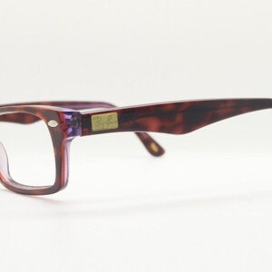 May include: A pair of tortoiseshell eyeglasses with purple tinted lenses. The glasses have a gold Ray-Ban logo on the side.