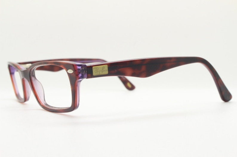 May include: A pair of tortoiseshell eyeglasses with purple tinted lenses. The glasses have a gold Ray-Ban logo on the side.