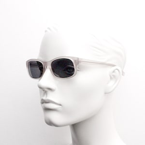 May include: Rectangular sunglasses with a translucent, light gray frame and dark lenses. The sunglasses are displayed on a white mannequin head, showcasing the eyewear's design and shape. The overall aesthetic is minimalist and modern.