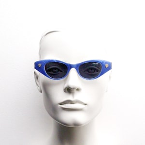 Op de afbeelding: Blauwe cat-eye zonnebril met een hartdetail aan elke kant. De zonnebril zit op een wit mannequins hoofd.