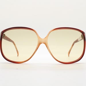 Op de afbeelding: Oversized vintage zonnebril met een bruin schildpadmontuur en geel getinte glazen. De bril heeft een grote, afgeronde vorm, met een brug die de twee glazen verbindt. De pootjes zijn dun en lopen door naar de oren.