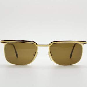 Peut inclure: Lunettes de soleil dorées avec des verres bruns et une bordure bordeaux. Les lunettes ont une forme carrée et un design classique.