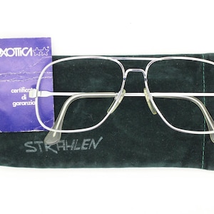 May include: Silver-framed eyeglasses with a classic aviator design, resting on a dark green case. The case has the word "STRAHLEN" printed on it. A blue tag with the word "LUXOTTICA" is also present.