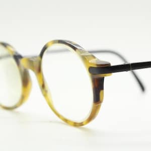 Könnte beinhalten: Runde Brille mit Schildpattmuster auf den Fassungen und schwarzen Bügeln. Die Gläser sind klar, und die Fassungen haben ein klassisches, Vintage-Design. Die Brille ist vor einem weißen Hintergrund platziert.