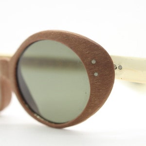 Może przedstawiać: Zbliżenie na vintage okulary przeciwsłoneczne z brązową, teksturowaną oprawką i zielonymi soczewkami. Zausszniki są w kolorze złamanej bieli. Okulary mają owalny kształt retro, na białym tle.