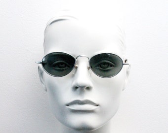 May include: A white mannequin head wearing oval-shaped sunglasses with a silver frame and dark gray lenses.