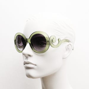 May include: Oversized round sunglasses featuring translucent olive green plastic frames with dark gradient lenses. The temples are designed with a distinct swirling scroll pattern that extends from the outer edges of the circular rims.