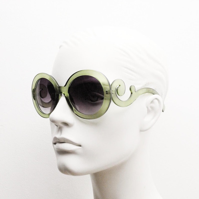 May include: Oversized round sunglasses featuring translucent olive green plastic frames with dark gradient lenses. The temples are designed with a distinct swirling scroll pattern that extends from the outer edges of the circular rims.