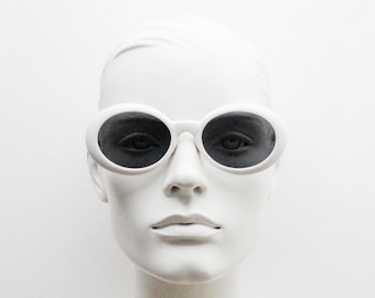 Puede incluir: Gafas de sol ovaladas blancas con lentes negros. Las gafas están en una cabeza de maniquí blanca.