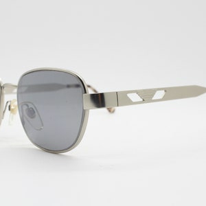 May include: Silver-toned sunglasses with gray lenses. The frames have a rectangular shape with a subtle geometric design on the temples. The earpieces feature a tortoiseshell pattern. A classic eyewear accessory.