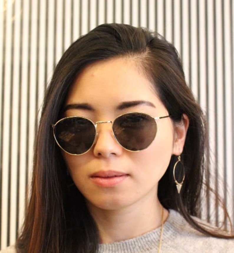 Pu&ograve; includere: Occhiali da sole rotondi con montatura dorata e lenti scure. Gli occhiali hanno una sottile montatura dorata. Lo sfondo &egrave; un motivo a strisce bianche e nere.