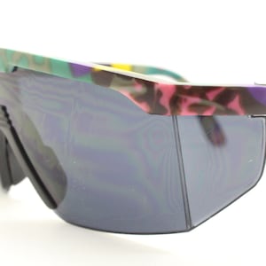 May include: Close-up of a pair of retro sunglasses with a shield-style lens. The frame features a colorful camouflage pattern in shades of green, pink, purple, and brown. The lenses are a dark gray color.