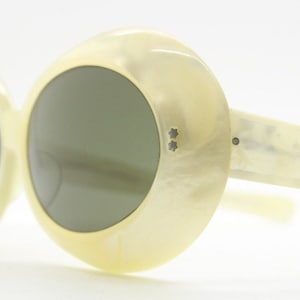 Może przedstawiać: Okulary przeciwsłoneczne w białym kolorze, okrągłe, o dużym rozmiarze, z ciemnymi szkłami. Okulary mają unikalne, fakturowane, perłowe wykończenie.