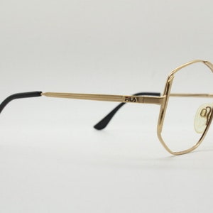Puede incluir: Gafas de metal doradas con patillas negras. La montura tiene una forma geom&eacute;trica y el nombre de la marca "FILA" est&aacute; impreso en el puente.