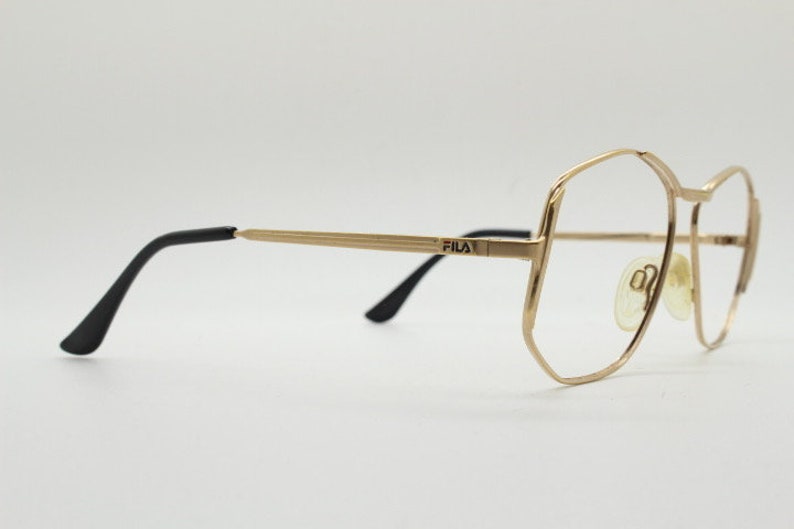Puede incluir: Gafas de metal doradas con patillas negras. La montura tiene una forma geom&eacute;trica y el nombre de la marca "FILA" est&aacute; impreso en el puente.