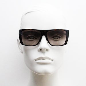 May include: Rectangular sunglasses featuring a dark brown tortoiseshell frame and gradient tinted lenses. The eyewear has a wide bridge and thick temples, providing a full-coverage look. The lenses transition from a darker shade at the top to a lighter tone at the bottom, set against a neutral white mannequin head.