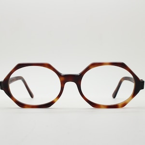 Puede incluir: Gafas con una forma de montura hexagonal distintiva que presenta un patrón clásico de carey en tonos marrón y beige. La montura tiene un acabado liso y pulido con un puente que conecta los dos cristales y varillas finas en los laterales.