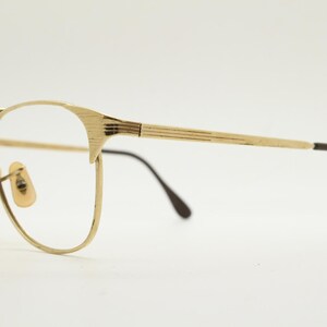 Puede incluir: Gafas de metal dorado con patillas de pl&aacute;stico marr&oacute;n. Las gafas tienen una forma rectangular redondeada.