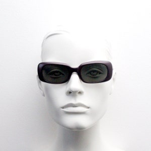 May include: A mannequin wearing a pair of dark purple rectangular sunglasses with black lenses.