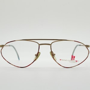 May include: Gold-toned aviator eyeglasses with a red and brown patterned rim. The glasses have clear lenses and a double bridge. The brand name "TREND COMPANY" is printed on the right lens.