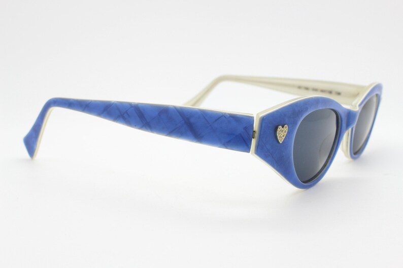 Op de afbeelding: Blauwe cat-eye zonnebril met een wit montuur en een klein hartvormig detail aan de zijkant.