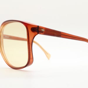 Op de afbeelding: Vintage oversized zonnebril met een warm amberkleurig montuur en geel getinte glazen. De monturen hebben een afgeronde, hoekige vorm en de veren zijn een donkerdere tint amber. Een klassiek brillenaccessoire.