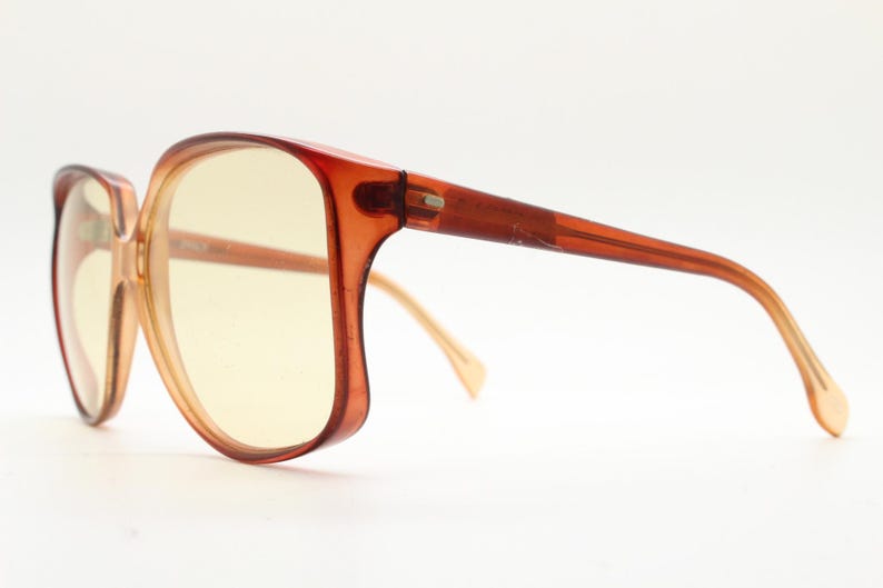 Op de afbeelding: Vintage oversized zonnebril met een warm amberkleurig montuur en geel getinte glazen. De monturen hebben een afgeronde, hoekige vorm en de veren zijn een donkerdere tint amber. Een klassiek brillenaccessoire.