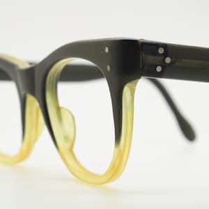 Op de afbeelding: Close-up van een vintage bril met een cat-eye vorm. De monturen zijn zwart met een geel getint interieur. De veren zijn donker en de glazen zijn helder. De bril heeft een retro esthetiek.