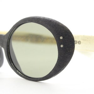 Może przedstawiać: Okulary przeciwsłoneczne w kształcie owalu, czarne, z fakturowaną oprawką i jasnozielonymi szkłami. Okulary mają białą plastikową zausznicę.