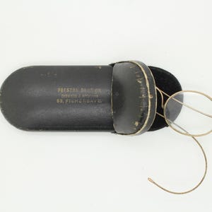 Peut inclure: Un étui à lunettes en cuir noir ouvert, contenant des lunettes rondes à monture dorée. L'étui porte l'inscription "PRESTON DAVIS CO. CHEVINS & WIFE 93, FISHERGATE". Les lunettes reposent sur un intérieur en velours noir.
