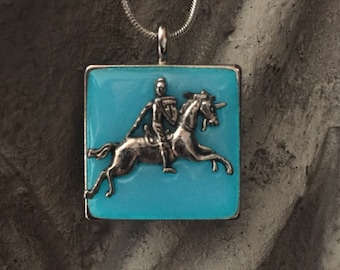 May include: Silver-toned pendant necklace featuring a square pendant with a light blue background. The pendant has a raised silver image of a knight on a horse. The necklace chain is a thin, silver snake chain.