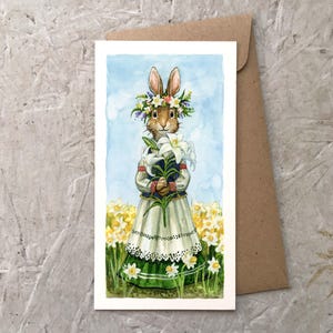 May include: Watercolor art print of a rabbit wearing a floral crown and holding white lilies. The rabbit is dressed in a traditional outfit, standing in a field of yellow flowers. The art print is on a white card with a brown envelope.