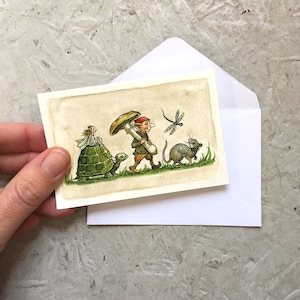 May include: A charming watercolour illustration on a card depicting a fairy on a turtle, a gnome with a mushroom, a dragonfly, and a mouse. The card is near a white envelope, suggesting a greeting card.