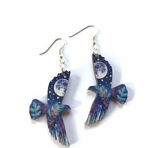 May include: A pair of wooden earrings featuring a blue and purple night sky with a full moon and stars. The earrings are shaped like a bird in flight.