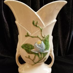 May include: A vintage ceramic vase with a pale pink glaze and a floral design. The vase has two handles and a delicate floral design featuring a blue flower with green leaves.