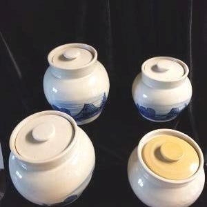 May include: Four ceramic jars with lids. Three are white with blue wave designs, and one has a yellow lid. The jars are round with a small knob on top of each lid. The jars are on a black surface.