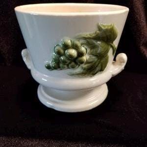 May include: White ceramic planter with a grapevine design. The planter has two handles and a pedestal base.