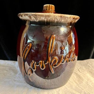 May include: A brown ceramic cookie jar with a glossy finish and a matching lid. The word Cookies is embossed in large, cursive script on the front of the jar. The rim of the jar and the lid feature a decorative drip-style glaze in cream and tan.