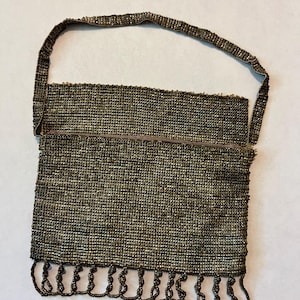 May include: A rectangular handbag covered in small, dark metallic beads. The bag features a single thin beaded strap attached to the top corners and a row of beaded loops hanging from the bottom edge, creating a textured, shimmering appearance.