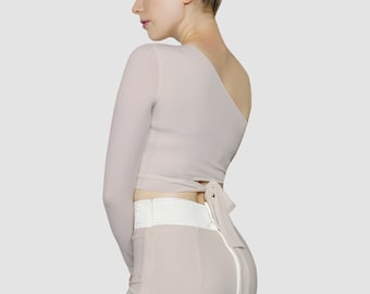 Op de afbeelding: Een tweedelige outfit in een lichtbeige kleur. Het topje is een langarmige, one-shoulder crop top met een strik aan de achterkant. De rok is een aansluitende midi-rok met een witte tailleband en een split aan de zijkant.