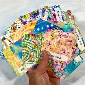 May include: A hand holds a fan of colorful cards with abstract designs and words like "hopeful" and "centered." The cards feature a variety of patterns, including watercolor-like washes, leaf prints, and geometric shapes. The color palette includes blues, yellows, and pinks.